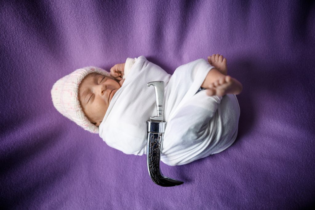 Newborn baby Elaahi with a family heirloom knife wrapped in a soft white wrap, lying on a purple backdrop during a baby photoshoot at Kirvan Photography + Film’s Thunder Bay studio.