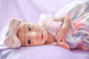 Close-up of newborn baby Elaahi’s face, wearing a flower dress, lying on a purple backdrop during a baby photoshoot at Kirvan Photography + Film’s Thunder Bay studio.