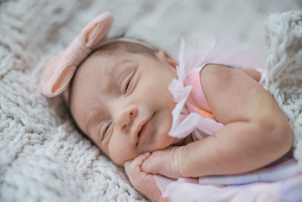Close-up of newborn baby Elaahi wrapped in a worn white blanket, hands together and smiling, resting on a purple backdrop during a baby photoshoot at Kirvan Photography + Film’s Thunder Bay studio.