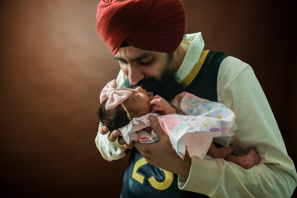 Father kissing newborn baby Elaahi, held gently in his arms on a soft brown backdrop during a family baby photoshoot at Kirvan Photography + Film’s Thunder Bay studio.