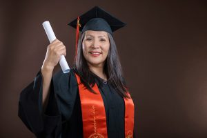 Older graduate in cap and gown, posing on a soft brown backdrop during a graduation photoshoot at Kirvan Photography + Film’s Thunder Bay studio.