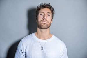 Close-up of Josh’s face in a white t-shirt and silver necklace, with bright lighting and a soft shadow cast behind him on a dark background.