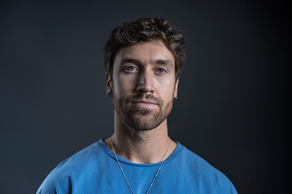 Josh wears a blue shirt under Rembrandt lighting, with part of his face in shadow and a triangle of light under one eye, set against a dark backdrop.