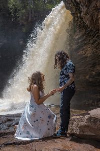 Sam smiles while on one knee, holding the ring, proposing to Jess at Wolf River Falls.