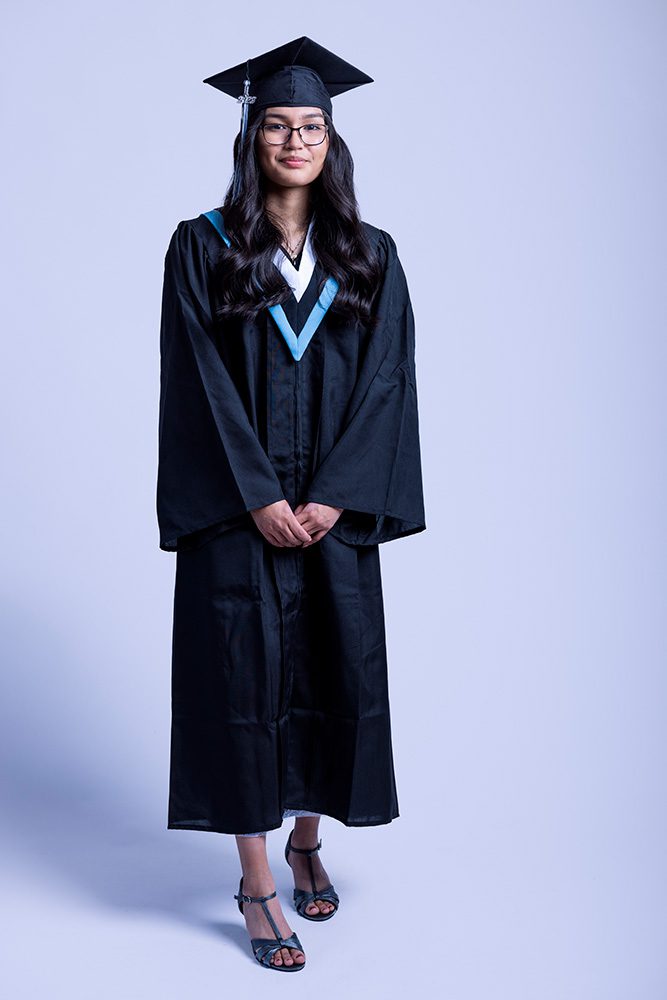 Full-body graduation photo of young Indigenous girl in cap and gown on white backdrop