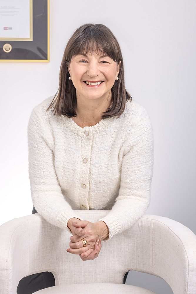Audrey from Royal LePage wearing a white sweater, leaning over a high-top chair and smiling confidently at the camera.