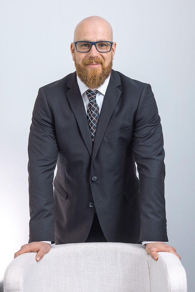 Jonathan from Royal LePage smiling at the camera while leaning on a high-top chair in a black suit.