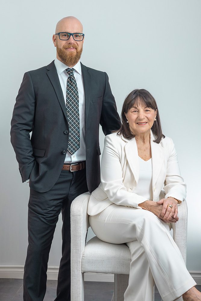Audrey seated in a white suit and Jonathan standing behind in a black suit, both smiling—professional real estate headshot.