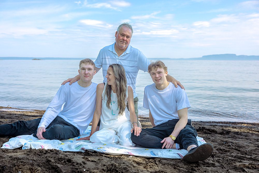 Sarah, her husband, and their two sons sit together on a blanket at the beach in front of Lake Superior, captured during their family session with Photography Gift Cards in Thunder Bay.