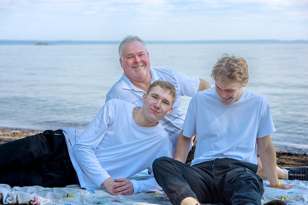 Sarah’s husband sits on a blanket at the beach with their two sons, smiling together with Lake Superior in the background—captured through Photography Gift Cards in Thunder Bay.