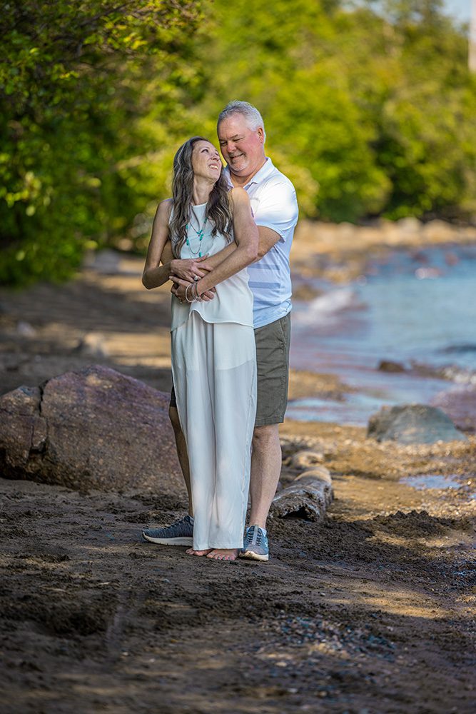 Sarah stands on the beach as her husband wraps her in a loving hug from behind; the shoreline stretches out behind them in this heartfelt moment captured through Photography Gift Cards in Thunder Bay.