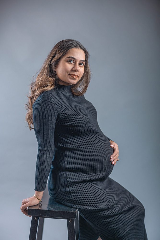 Ayush seated on a stool during her Studio Maternity Photography session, resting one hand on her baby bump and looking confidently at the camera.