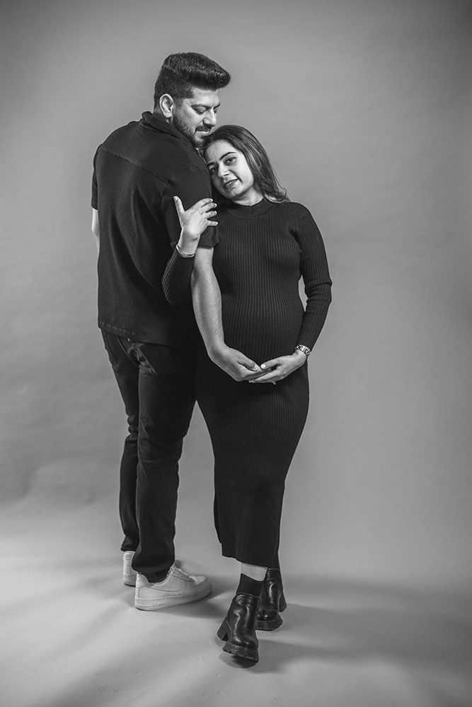 Full-body portrait of Ayush leaning on her husband’s arm and looking into the camera during their Studio Maternity Photography session in Thunder Bay.