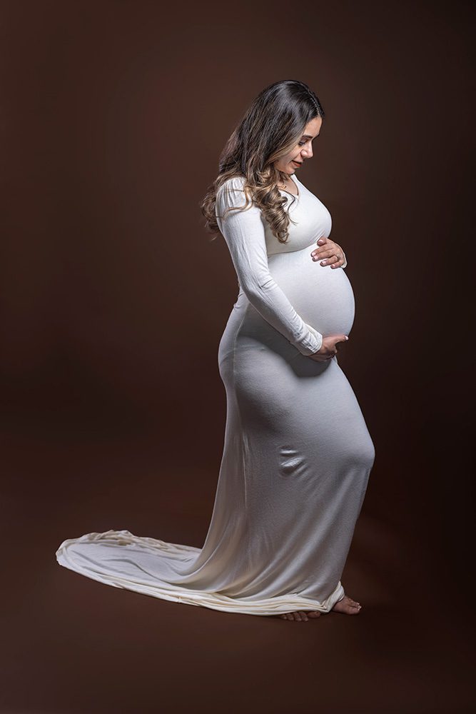 Ayush in a white dress on a brown backdrop, looking at her baby bump with wonder and joy during her Studio Maternity Photography session in Thunder Bay.