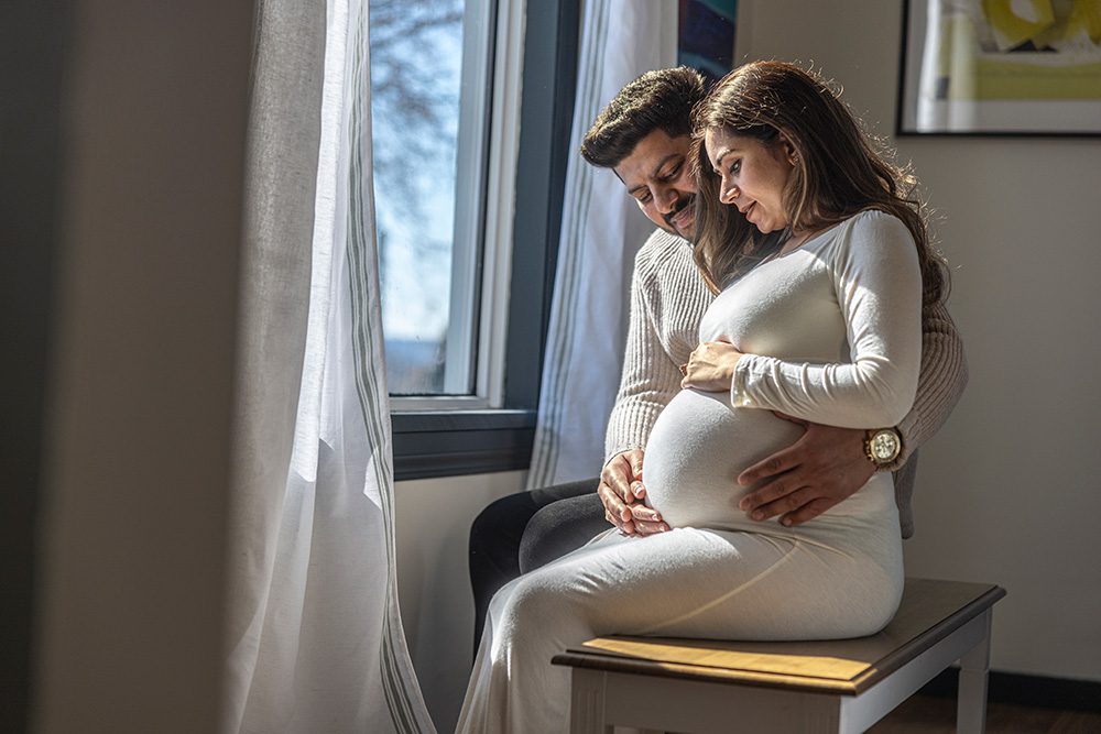 Ayush and her husband sit together in front of a window, sharing a candid, natural moment during their Studio Maternity Photography session in Thunder Bay.