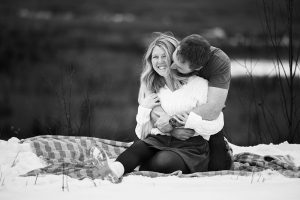 Steph smiles brightly as Mac hugs her from behind on a blanket near a snowy cliff during their winter engagement shoot in Thunder Bay with Kirvan Photography + Film.