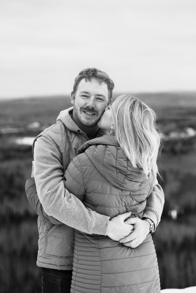 Black and white photo of Steph kissing Mac on the cheek during their winter engagement session in Thunder Bay with Kirvan Photography + Film.