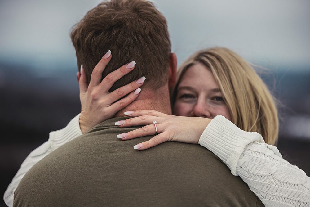 Steph peeks over Mac’s shoulder with her hands around his neck, showing her engagement ring during their winter engagement session in Thunder Bay with Kirvan Photography + Film.