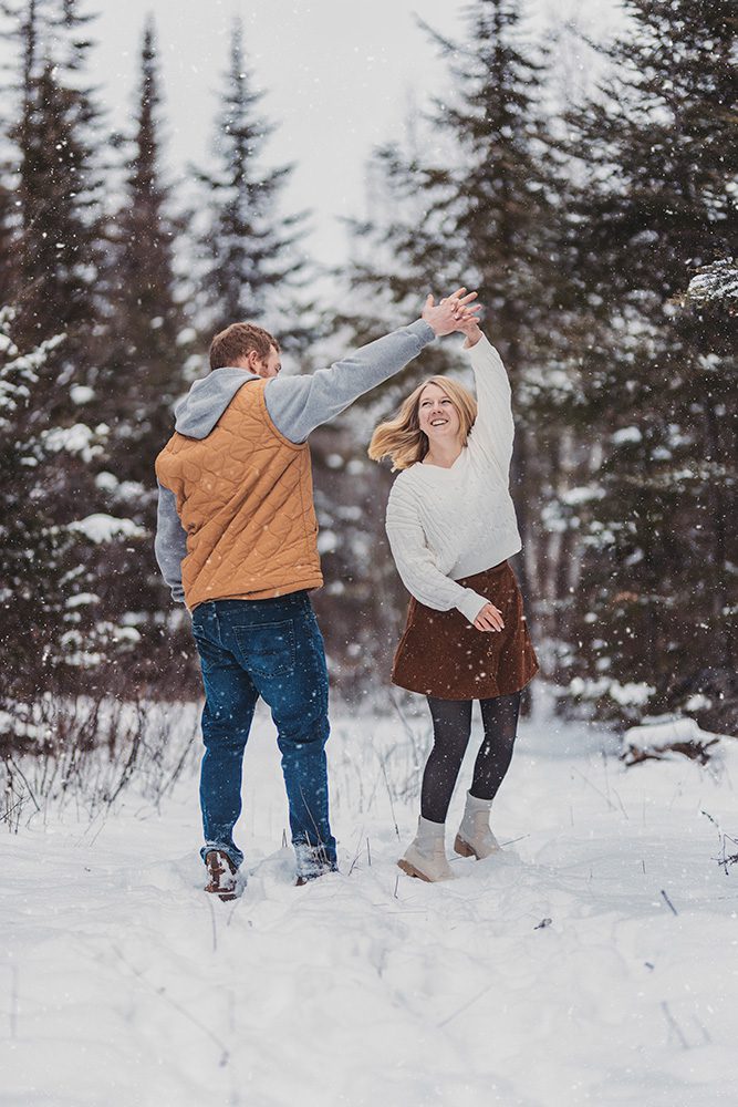 Mac and Steph hold hands high above as Steph twirls playfully beneath them during their winter engagement shoot in a snow-covered Thunder Bay forest.