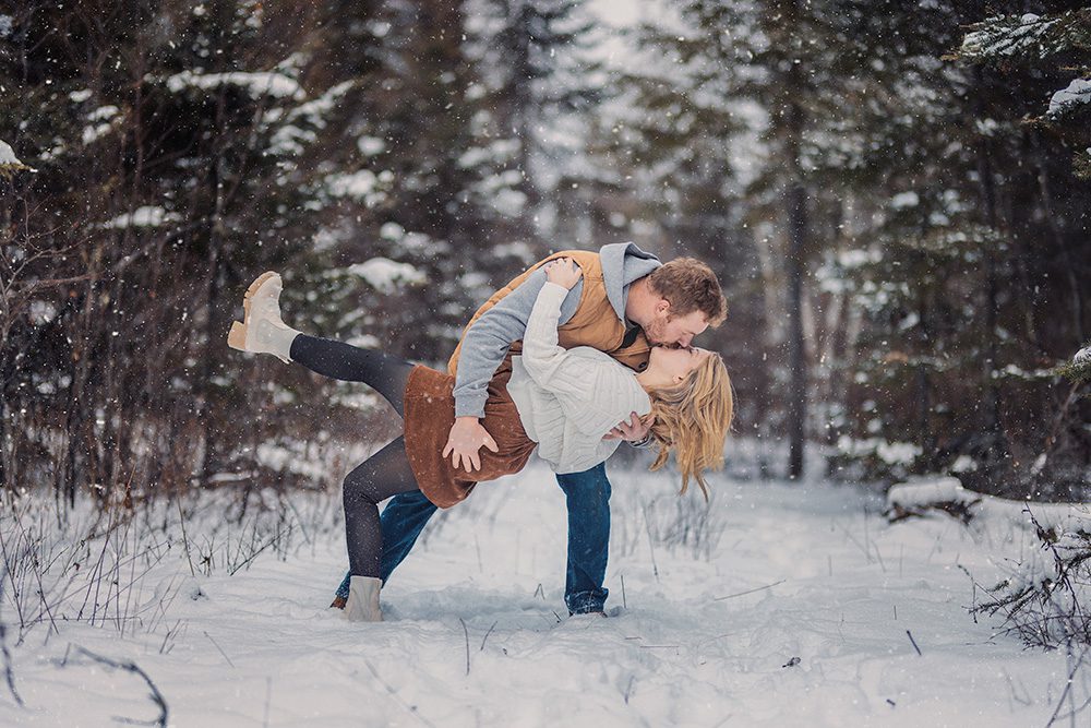 Mac dips Steph into a dramatic kiss as soft snow falls around them in a winter engagement session