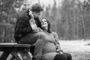 Sophie sits on the bench of a picnic table, looking up lovingly at Jeff as she reaches to touch his face; he sits on the tabletop above her while snow gently falls around them at Chippawa Park in Thunder Bay.