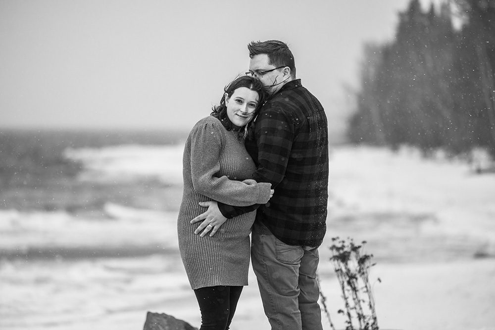 Black and white photo of Sophie and Jeff standing close together on a chilly, windswept beach at Chippawa Park in Thunder Bay, wrapped in the quiet drama of the moment.