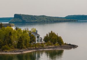 Modern lakefront house on rocky shoreline with forested hills and calm water reflections