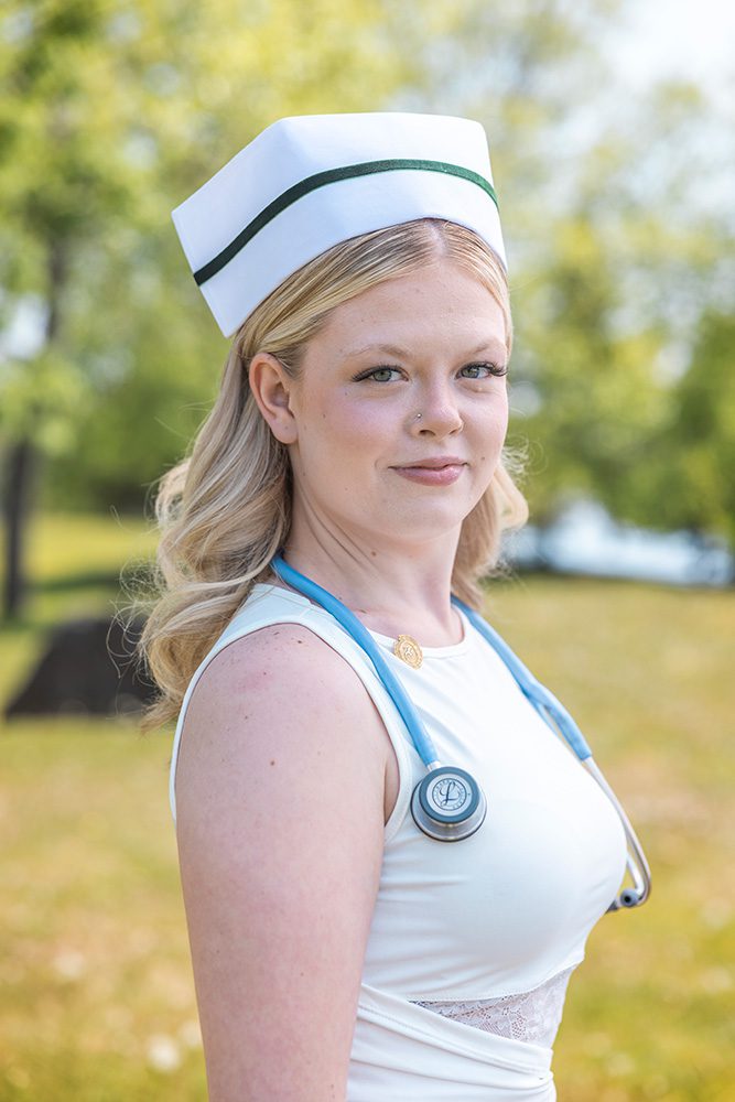 A young blonde woman in nursing uniform wearing a white nurse cap with green trim and a stethoscope around her neck, smiling confidently at the camera in an outdoor park setting with blurred green foliage background