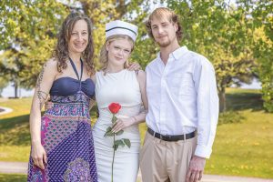 Graduate in white nursing outfit holding a rose, standing with her mother and boyfriend in a sunny outdoor park setting.