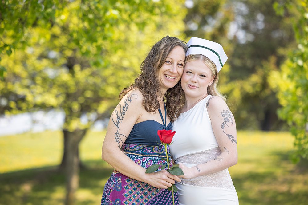 Two women embracing outdoors in a park setting, one wearing a colorful strapless dress holding a red rose, the other in white clothing with a white hat, both smiling at the camera with green trees in the background