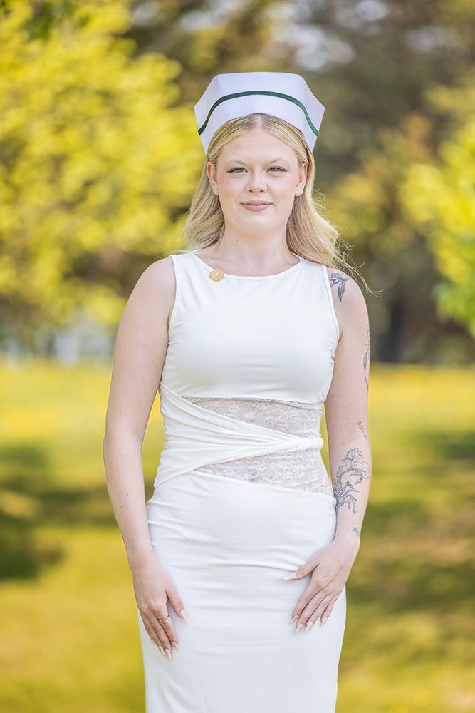 Young woman in a white nursing outfit and cap, standing confidently outdoors with trees and sunlight in the background.