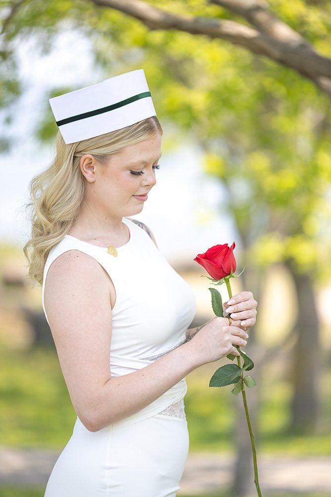 A young blonde woman in white nursing uniform and traditional nurse cap looking down thoughtfully at a single red rose she's holding, standing in a park with blurred green trees in the background