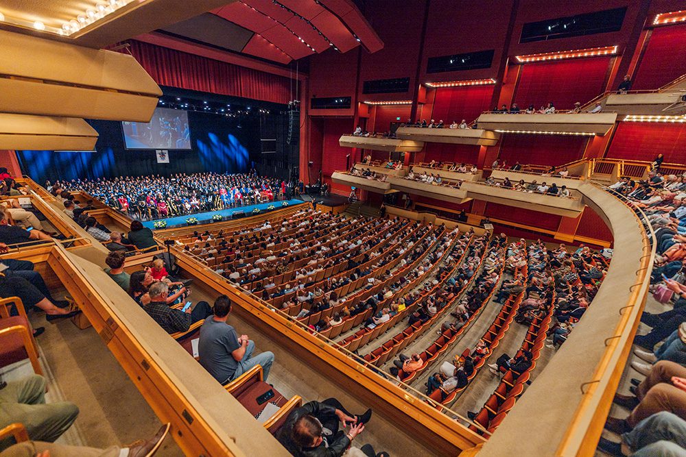 Wide-angle view of a large indoor graduation ceremony with graduates on stage and a full audience seated in a theatre-style auditorium.