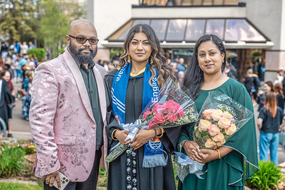 Graduate in cap and gown with blue ceremonial stole standing between two family members holding flower bouquets on campus with crowds in background