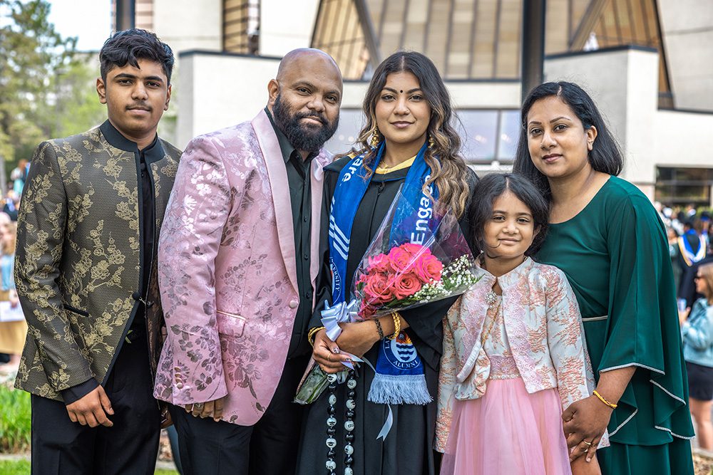 Graduate in gown with blue ceremonial stole holding pink roses surrounded by five family members in traditional and formal attire on campus