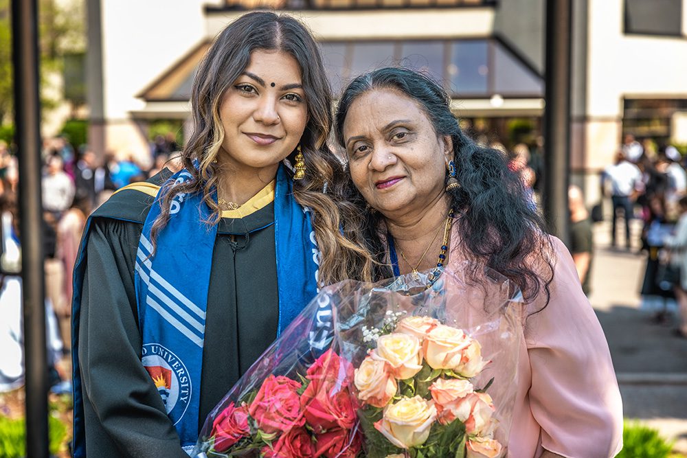 Graduate in cap and gown with blue ceremonial stole posing with woman in pink outfit holding colorful flower bouquet on campus