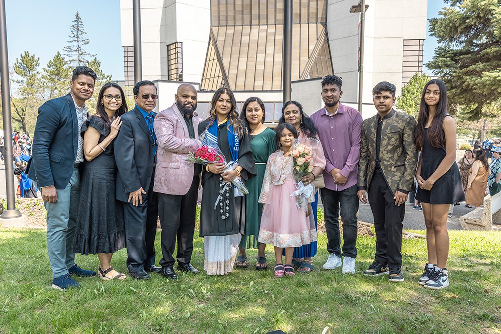 Large group of eleven family members and friends posing with graduate in gown and blue ceremonial stole holding flower bouquets in front of modern university building