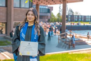 Smiling graduate in cap and gown holding diploma on university campus with modern buildings and people in background