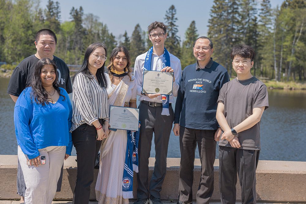 Group of seven people including friends and family posing by a lake with two graduates holding degree certificates, one in traditional sari and one in business attire with blue ceremonial stole