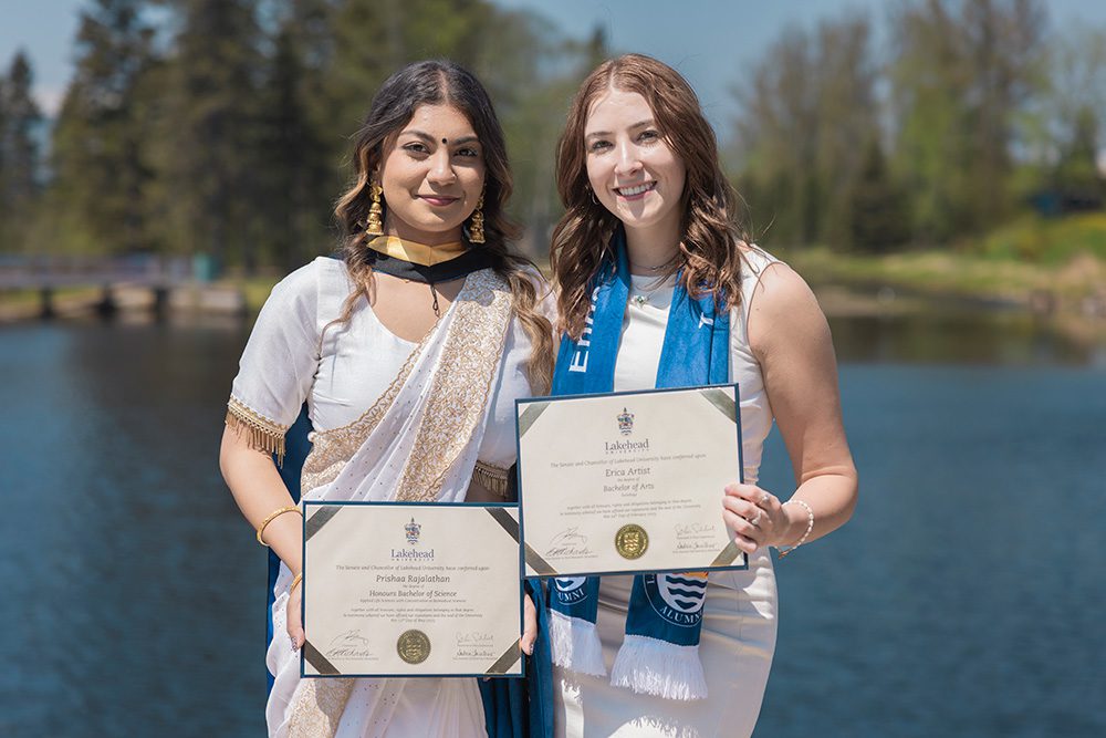 Two female graduates smiling and holding their degree certificates by a lake, one in white sari with gold jewelry, the other in white dress with blue ceremonial stole