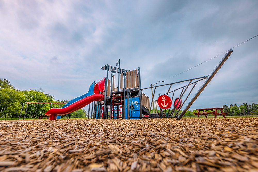 Pirate-themed playground with climbing structure, slides, and ship mast on wood chip surface with cloudy sky background