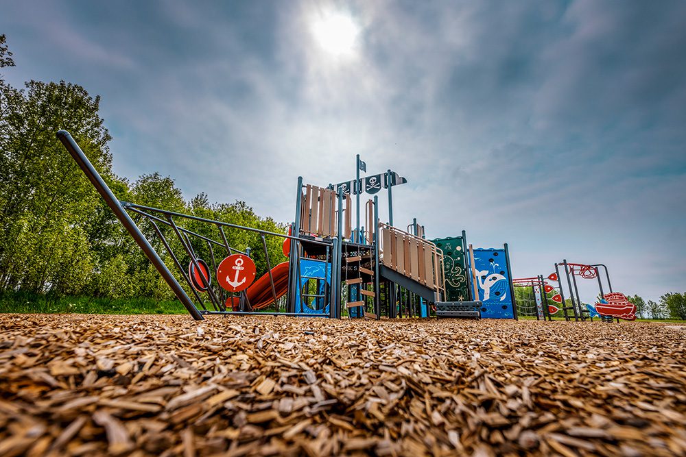 Wide view of pirate ship playground with climbing structures, slides, and ship mast on wood chip ground with trees in background
