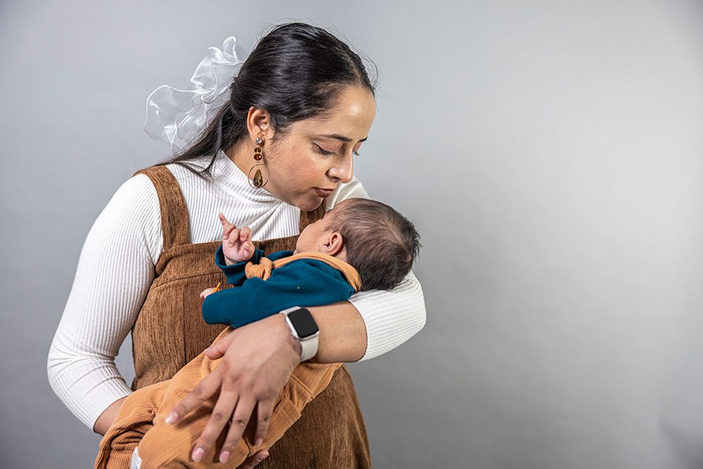 Woman with dark hair in white ribbed sweater and brown overalls gently kisses baby's head while holding infant against light gray background