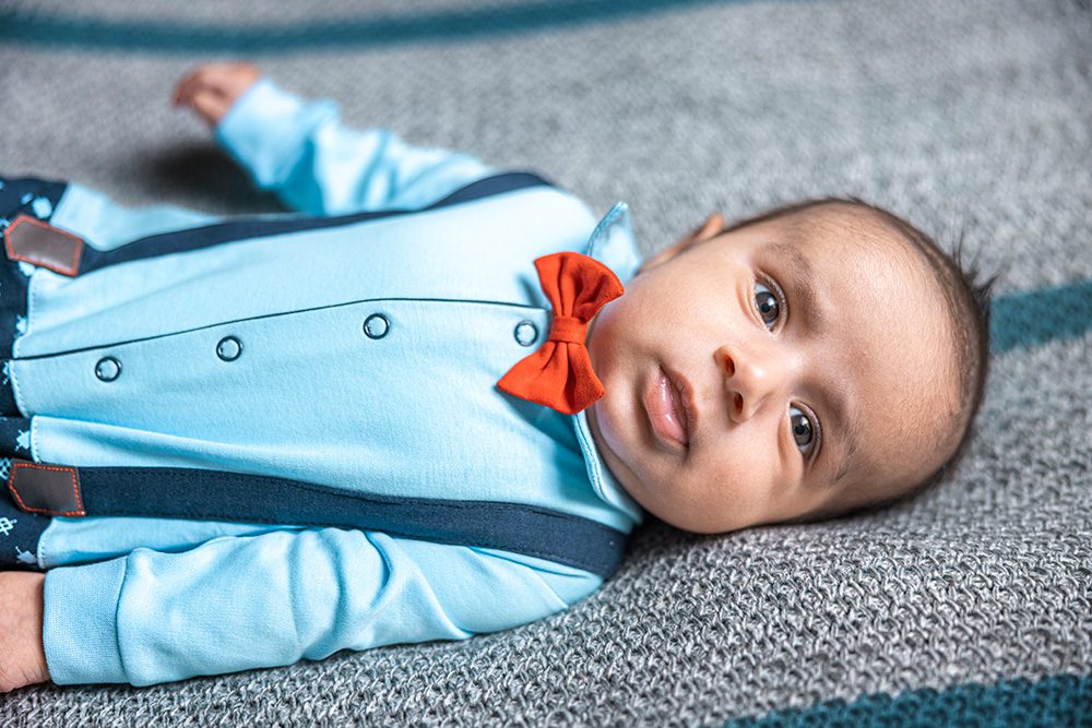Baby lying on a textured gray and blue blanket wearing a light blue outfit with dark suspenders and a red bow tie, looking directly at the camera.