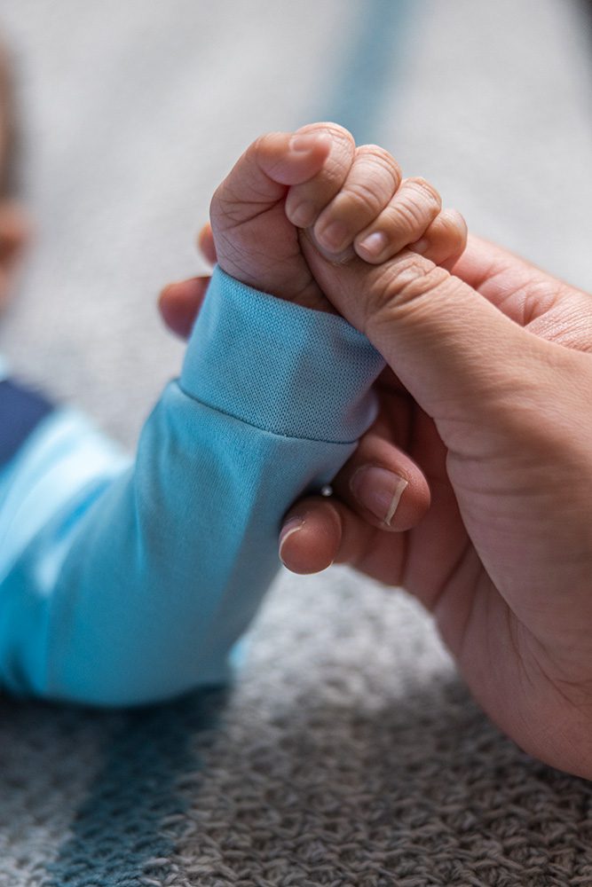 Close-up of adult hand gently holding baby's tiny clenched fist in blue sleeve against soft blurred background