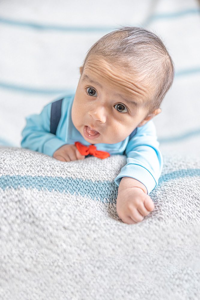 Baby in a light blue outfit with a red bow tie lying on a soft gray and blue blanket, looking up with a curious expression.