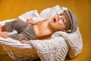 Baby wearing glasses and vintage-style outfit lying in a white basket on a cozy blanket.