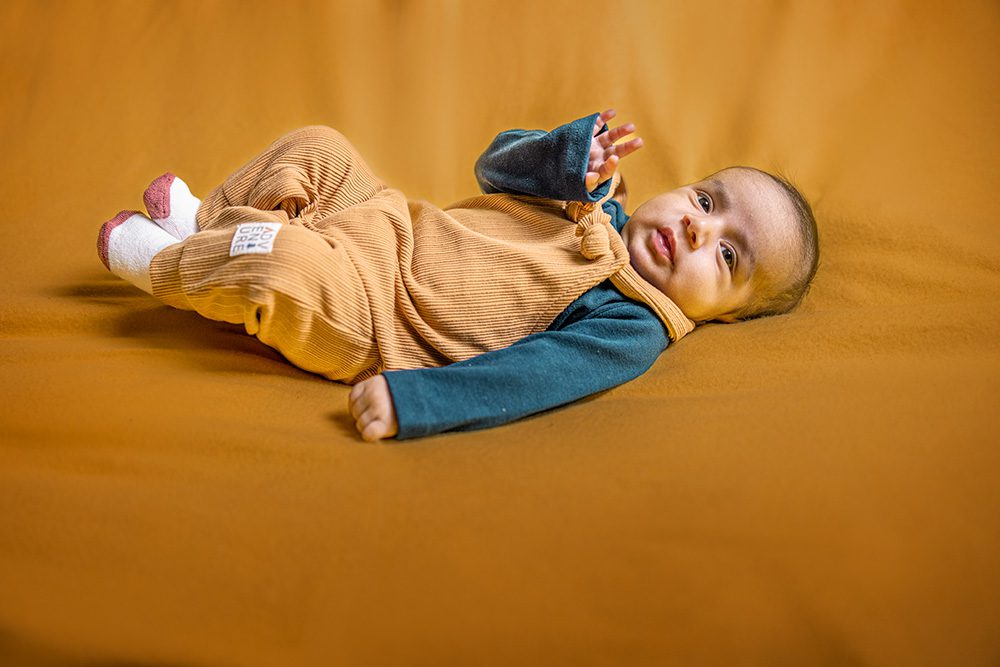 Baby wearing tan corduroy overalls and teal long-sleeve shirt lies on orange background with white socks with pink accents