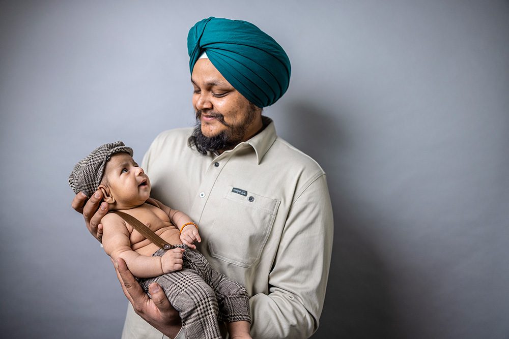 Man wearing teal turban and beige polo shirt holds baby dressed in brown checkered outfit and matching hat against gray background