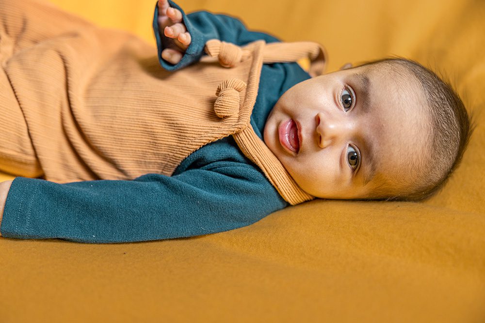 Baby wearing a teal ribbed sweater and tan overalls lies on orange background looking directly at camera with alert expression